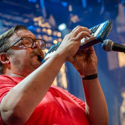 Das Bild zeigt einen Mann im roten Oberteil, welcher auf einer Melodica spielt.