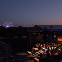 Das Bild zeigt eine Aufnahme der Bühne von oben. Es ist bereits dunkel. Das Orchester ist jecoh hell beleuchtet. Im Hintergrund sieht man beleuchtete Fahrgeschäfte des Volksfestes.