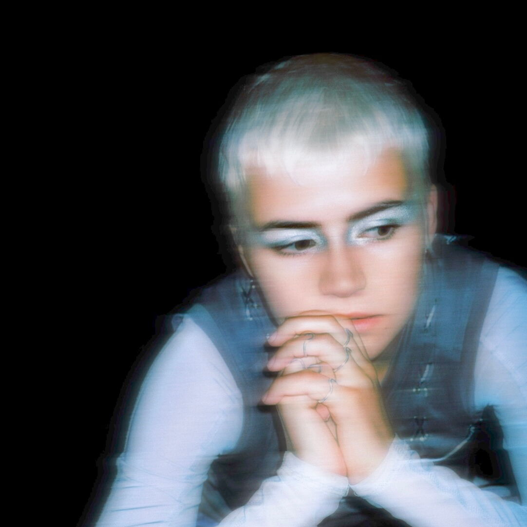 Das Bild zeigt Véra Marie Deubner aus dem Musikprojekt Epilog. Das Foto zeigt nur ihren Oberkörper, sie hat ihre Hände vor ihrem Kinn zusammengefaltet. Der Hintergrund ist schwarz und das Bild ist leicht verschwommen. Sie hat kurze weiße Haare, einen auff