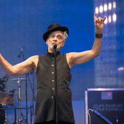 Das Bild zeigt Melingo am Mikrofon. Er streckt die Arme nach oben, wobei er in der rechten Hand ein Instrument hält.