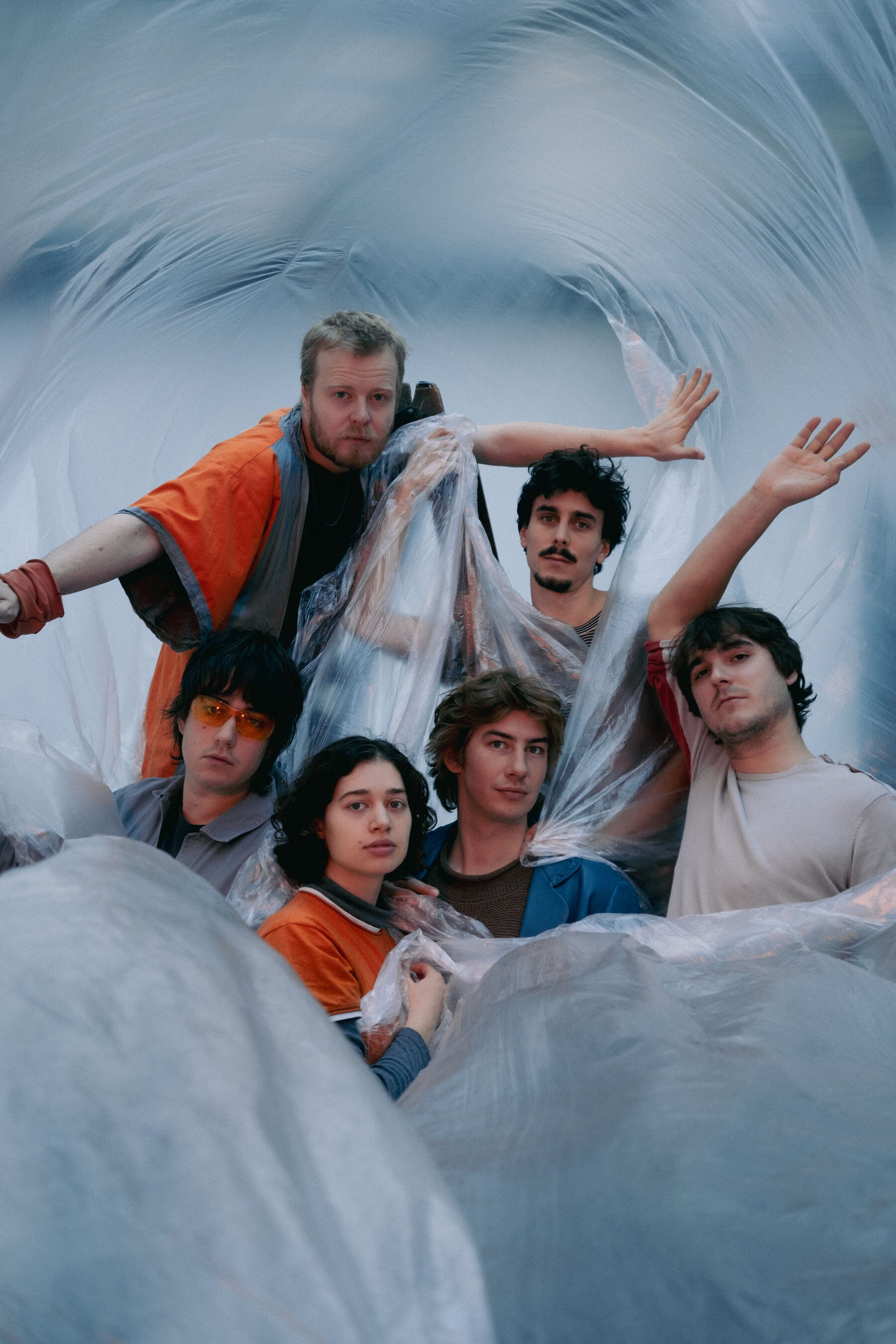 Das Bild zeigt 5 Personen, welche eng aufeinander sitzen. Um sie herum ist durchsichtige Folie, mit welcher sie sich zum teil auch zudecken.