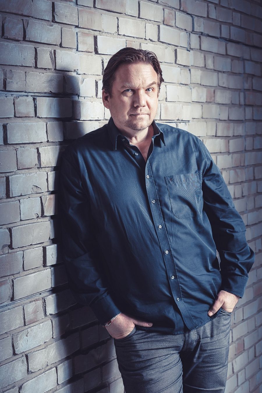 Matthias Brodowy lehnt lässig an einer grauen Backsteinmauer, die Hände in die Hosentaschen gesteckt und den Blick in die Kamera gerichtet. Er trägt ein blaues Hemd und wird von hellem Scheinwerferlicht bestrahlt.