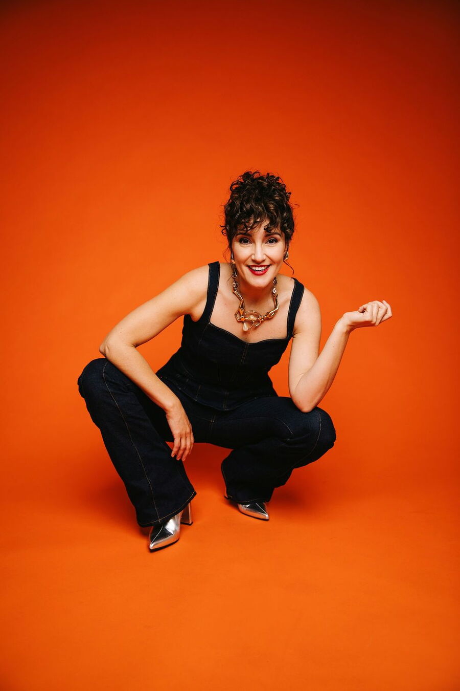 Ina Regen posiert vor einem leuchtend orangefarbenen Hintergrund. Sie trägt einen ärmellosen, dunklen Jumpsuit aus Jeansstoff, silberne spitze Schuhe und eine auffällige Kette mit großen Gliedern. Ihre hochgesteckten, lockigen Haare fallen in ihr Gesicht