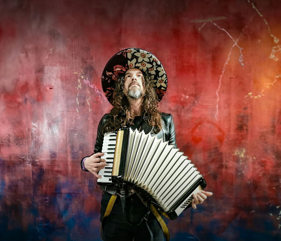 Das Bild zeigt Luca Bassanese, er hat sein Akkordeon umhängen. Er steht vor einem roten Hintergrund welcher einer von Rissen durchzogen ist. Er trägt einen großen, auffälligen Hut. Das Bild ist frontal aufgenommen, er selbst schaut jedoch nach oben.