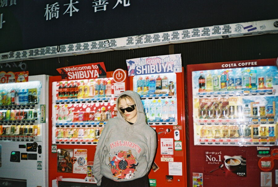 Das Bild zeigt die Sängerin Verifiziert vor einigen Getränke- und Snackautomaten aus Japan. Sie ist in der unteren Mitte des Bilds und trägt einen grauen Hoodie mit rotem Aufdruck, eine schwarze Sonnenbrille und sie hat blonde Haare. Das Bild scheint bei