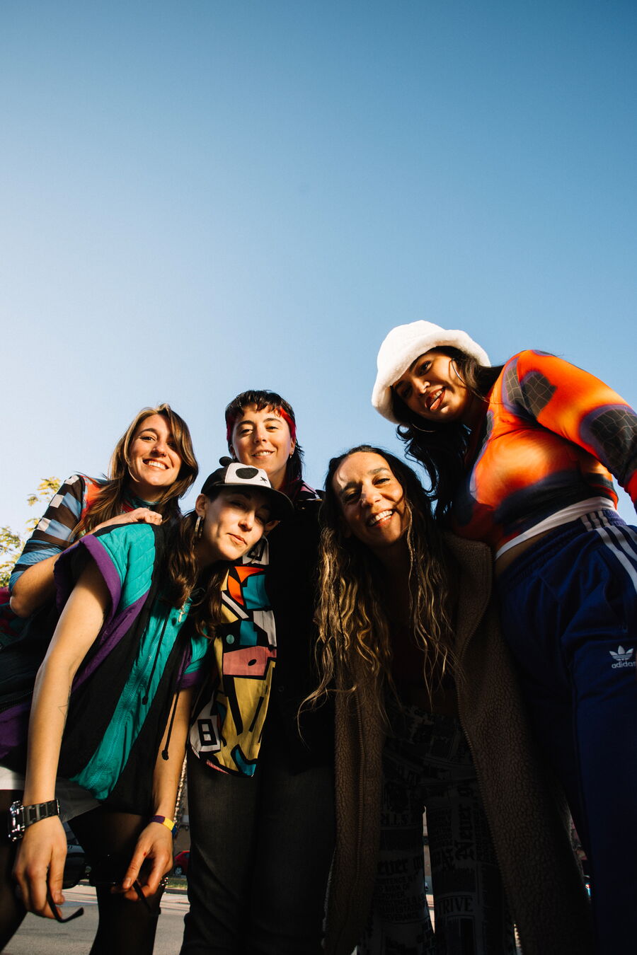 Das Bild zeigt die Fünf Mitglieder der Band Ketekalles. Das Foto ist in einer Froschperspektive von unten, unterm freien Himmel, aufgenommen worden. Sie lächeln alle in die Kamera und gepaart mit ihrer bunten, knalligen Kleidung mit verschiedenartigen Mus