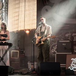 Zwei Musiker auf der Bühne, links eine Frau am Keyboar, rechts ein Mann an der Gitarre. Beide haben ein Mikrofon vor sich stehen und singen. Das Bild ist seitlich aufgenommen.