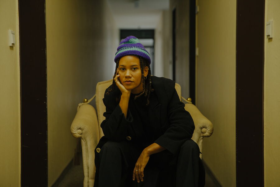 Das Bild zeigt Ami Warning in einem beige farbenen Gang auf einem weißen Sessel sitzen. Sie hat ihren Kopf auf ihre Hand gestutzt und schaut mit einem neutralen Blick in die Kamera.Sie ist in ganz schwarz gekleidet, bis auf ihre blau-lila gestreifte Mütze