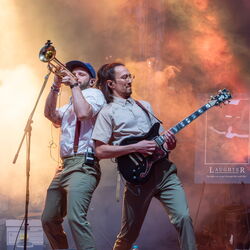 Auf dem Bild sind zwei Musiker in grünen Hosen welche sich nach hinten lehnen, sodass sich ihre Schultern berühren. Der Linke spielt Trompetem, der Rechte Gitarre.