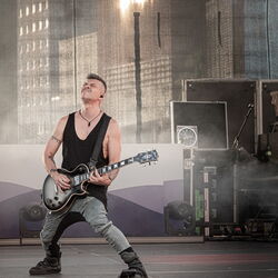 Ein Mann in grauer Hose und schwarzem Oberteil, welcher leidenschaftlich Gitarre spielt.