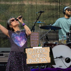 In der Mitte befindet sich ein Schild aus Pappe, auf welchem im weißer Schrift "Glitzer ist für alle da" steht. Links ist eine Sängerin, welche den rechten Arm hebt und mit dem Zeigefinger in die Luft zeigt. Rechts ist ein Schlagzeuger mit Sonnenbrille.